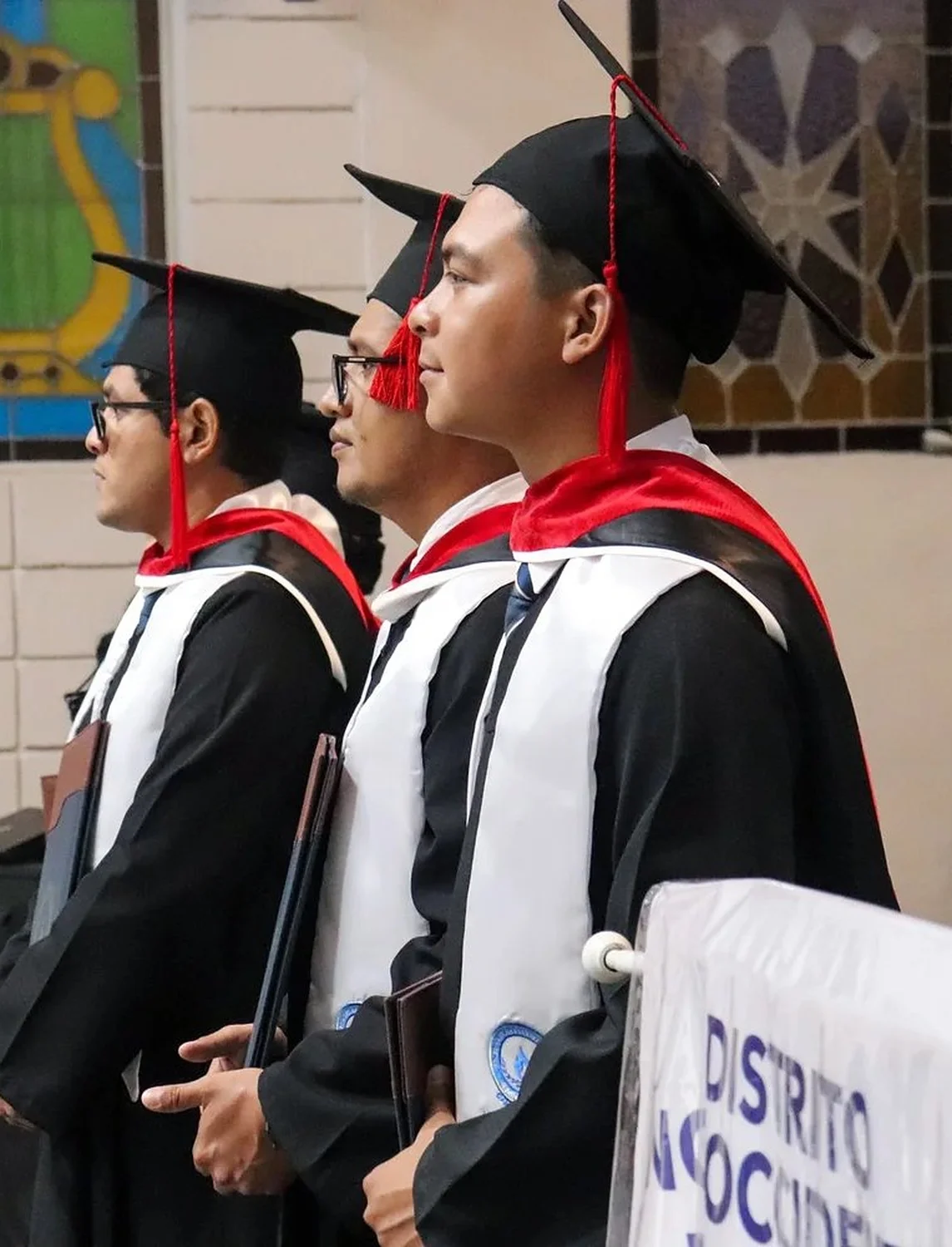 Tres graduandos del seminario con toga y birrete, de perfil, junto a la bandera del Distrito